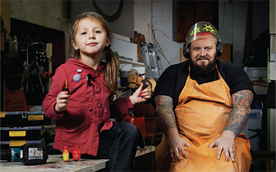 A father in an apron sits next to his daughter, who is painting his nails.