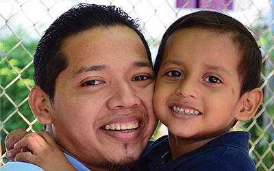 A father from Central America holds his young son, cheek to cheek.