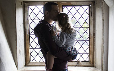 A father holding his daughter in front of a window kisses her on the forehead.