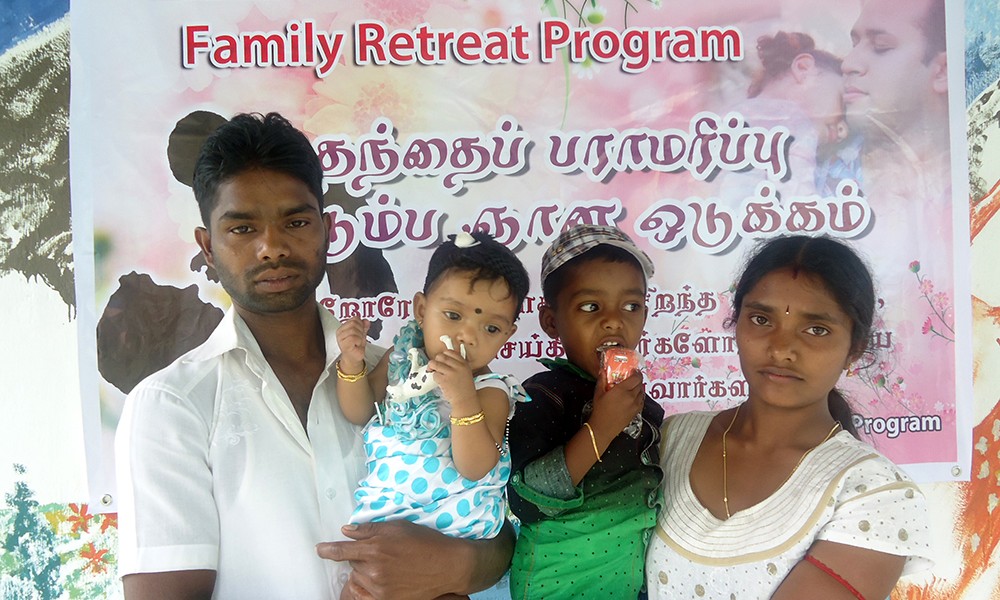 Ravichandran, a father in Sri Lanka, holds one of his children in his arms. His wife stands next to him holding their second child. In the background, a banner reads "MenCare Family Retreat Program."