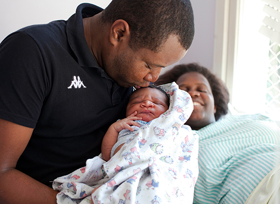 A father kisses his child in the hospital after birth, while his wife, who is lying down, smiles.